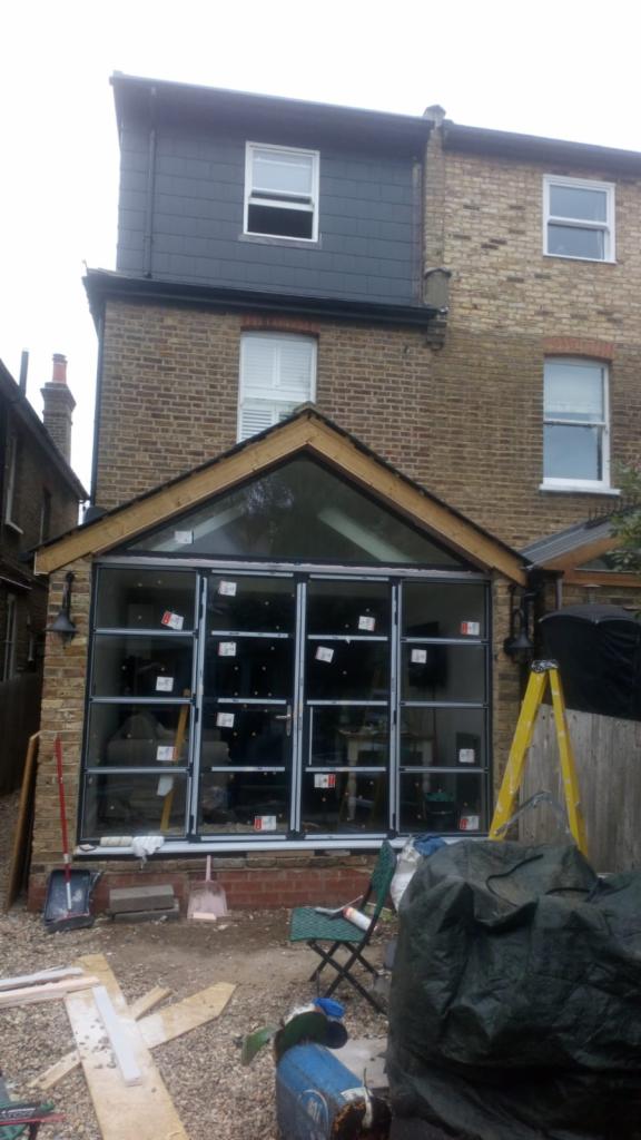 Rozbudowa domu z dużymi, przeszklonymi drzwiami przesuwnymi, widoczne prace wykończeniowe, drabina i narzędzia.