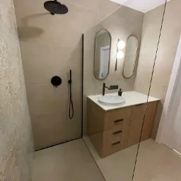 Nowoczesna łazienka z kabiną prysznicową typu walk-in, czarną armaturą, owalnymi lustrami z podświetleniem i szafką pod umywalką z drewnopodobnym wykończeniem.