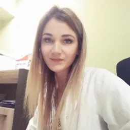 Portret uśmiechniętej kobiety w białym fartuchu lekarskim, z certyfikatem widocznym w tle.