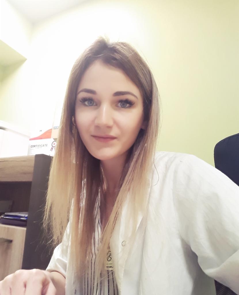 Portret uśmiechniętej kobiety w białym fartuchu lekarskim, z certyfikatem widocznym w tle.