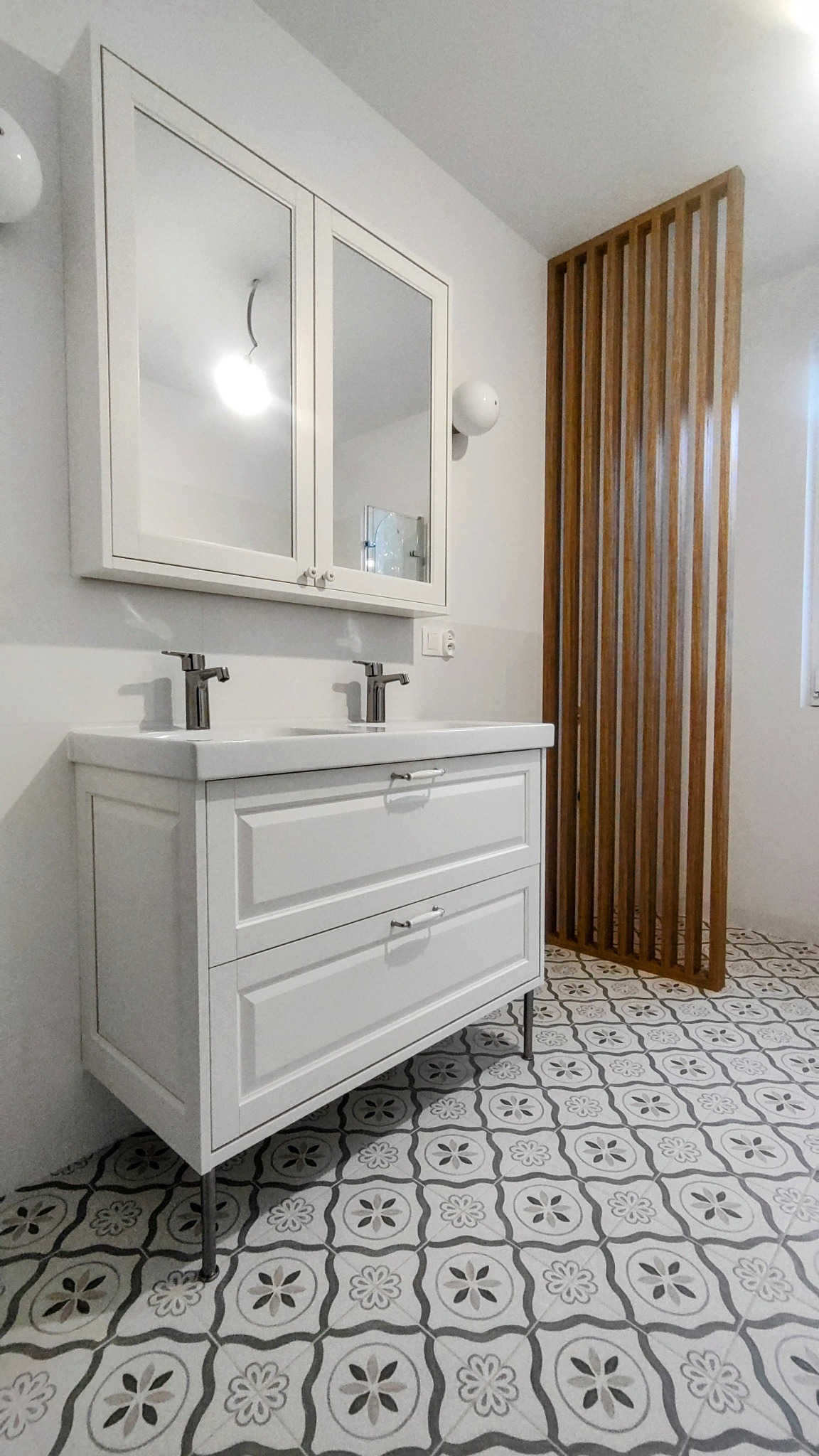 Biała szafka łazienkowa z umywalką i lustrem na tle wzorzystej podłogi i drewnianej ścianki działowej. Minimalistyczny design, jasne wnętrze.