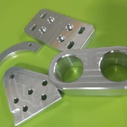 Cztery precyzyjnie obrobione, aluminiowe elementy o różnych kształtach, z widocznymi otworami i śladami obróbki, ułożone na jaskrawym, zielonym tle.