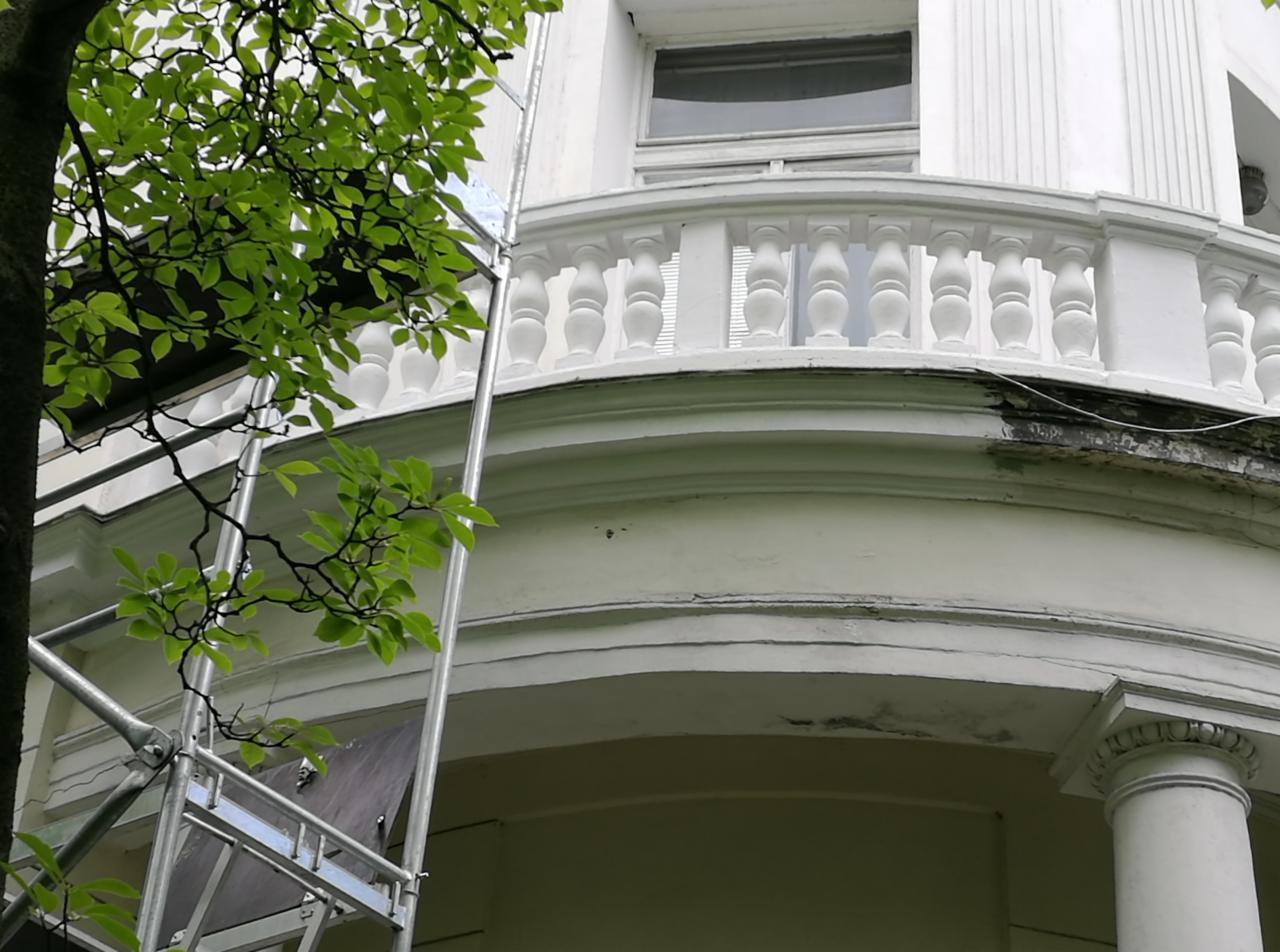 Biała, zaokrąglona elewacja budynku z balustradą i kolumnami, częściowo uszkodzona i przygotowywana do renowacji, widoczny fragment rusztowania i gałęzie drzewa z liśćmi.