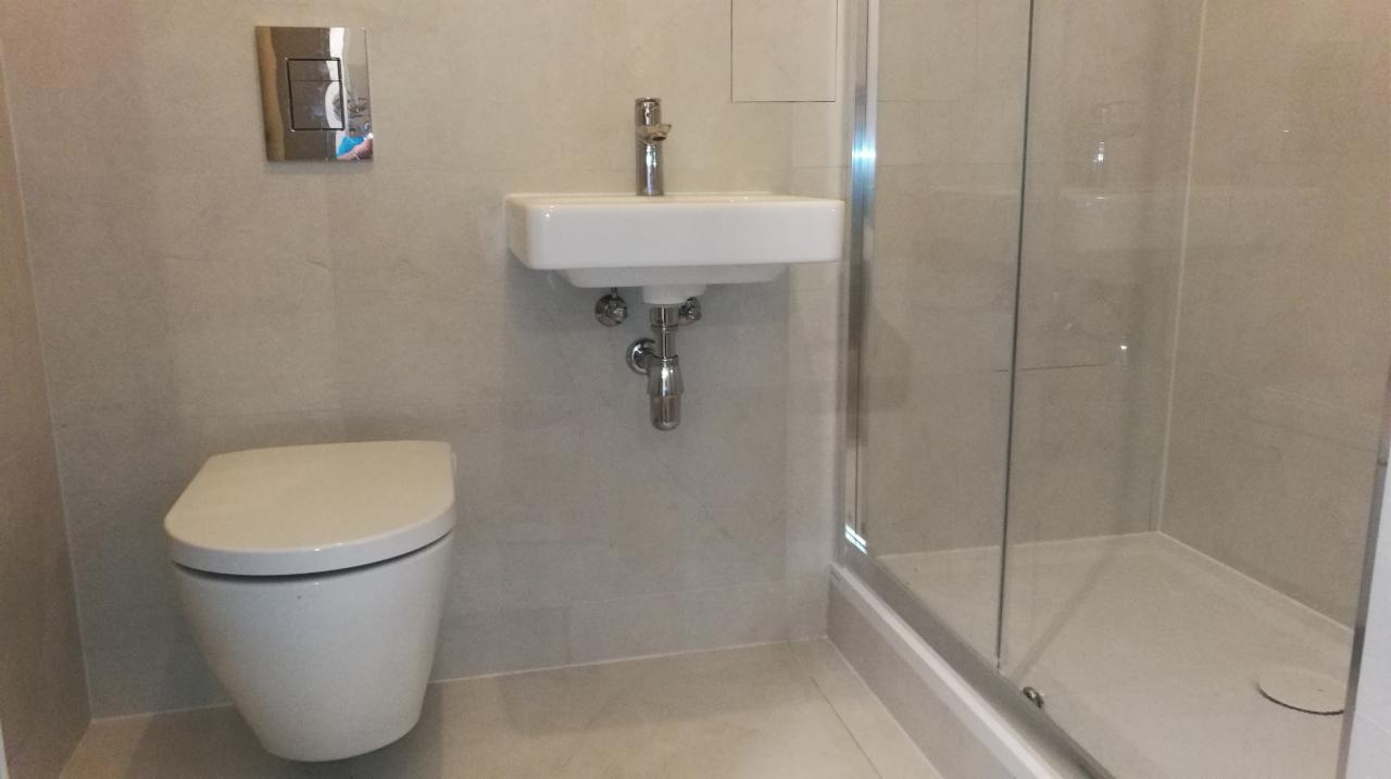 Nowoczesna łazienka z podwieszaną toaletą, małą umywalką i kabiną prysznicową z transparentnego szkła, wykończona jasnymi płytkami ceramicznymi.