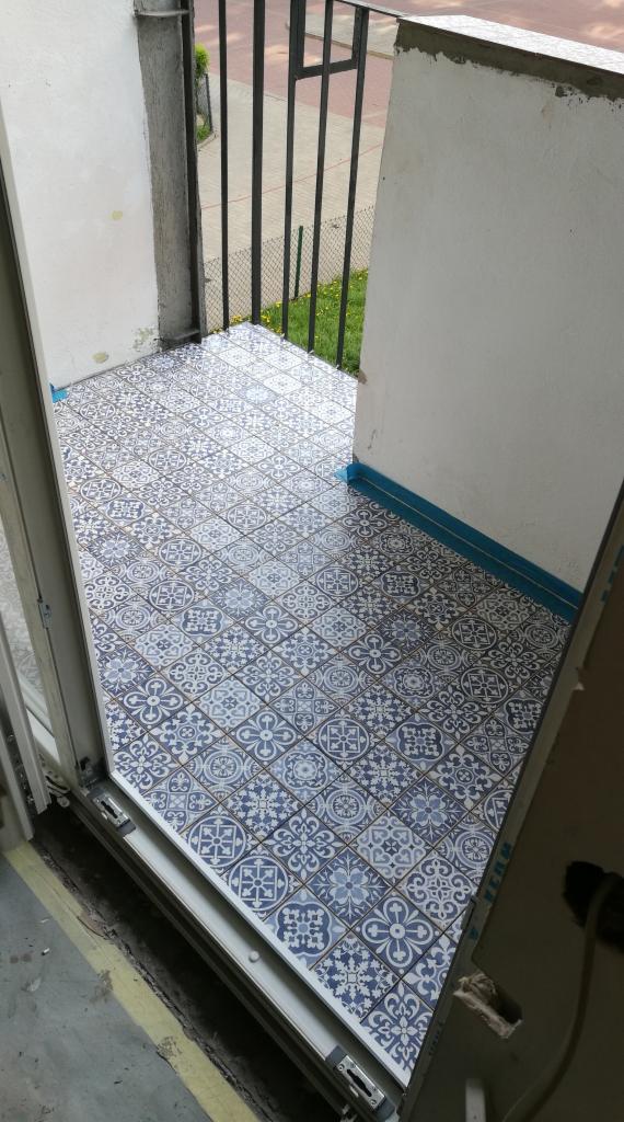 Balkon wyłożony wzorzystą, niebiesko-białą glazurą, widok z wnętrza mieszkania, z fragmentem ramy okiennej i widokiem na zieleń za barierką balkonu.