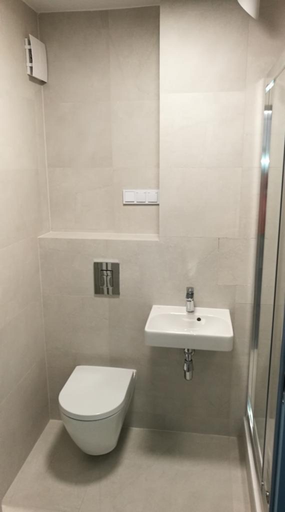 Nowoczesna, mała łazienka wykończona jasnymi, matowymi płytkami, z podwieszaną toaletą, umywalką oraz fragmentem kabiny prysznicowej.