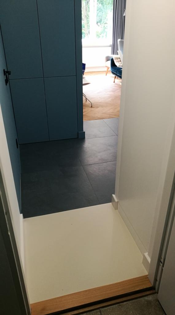 Wnętrze z korytarzem wykończonym ciemnymi, matowymi płytkami podłogowymi, białą podestem i widoczną częścią szafy w kolorze niebieskim; w tle widać salon z parkietem i meblami.