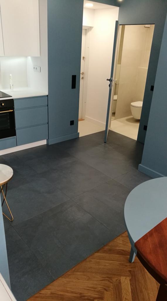 Nowoczesna łazienka z widoczną toaletą, połączona z kuchnią poprzez otwarte drzwi, na podłodze szara, kwadratowa glazura, w tle fragment parkietu ułożonego we wzór jodełki.