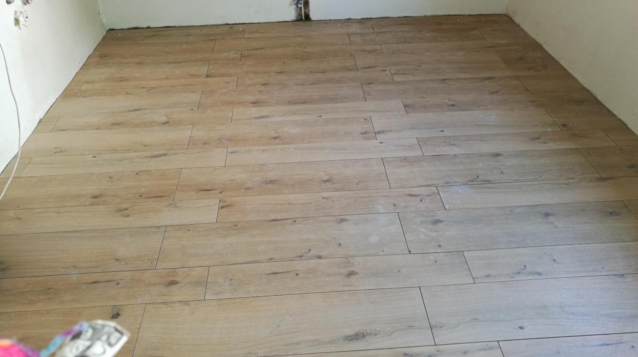 Ułożona podłoga z paneli imitujących drewno w jasnym odcieniu, widoczna w narożniku pomieszczenia z białymi ścianami i wystającymi rurami.