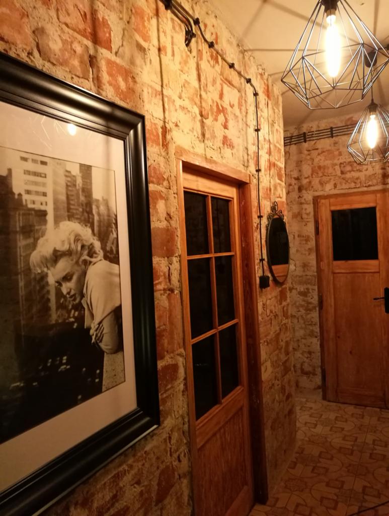 Korytarz z ceglanymi ścianami, drewnianymi drzwiami, czarno-białym portretem w ramie i geometrycznymi lampami wiszącymi.