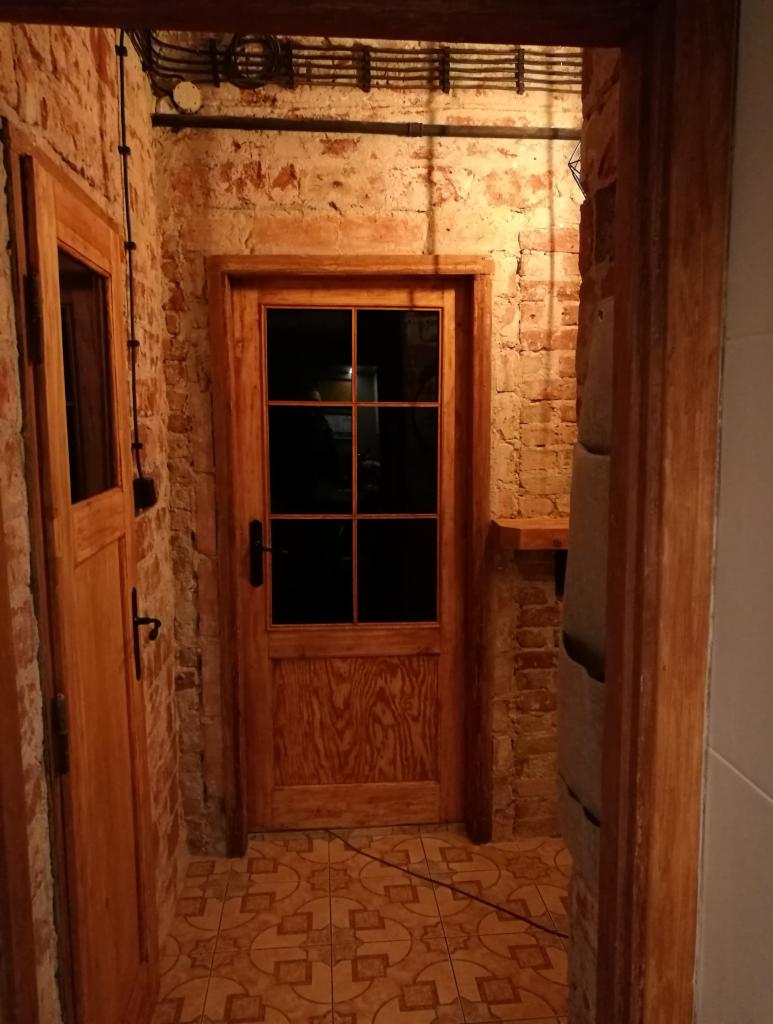 Korytarz z dwiema drewnianymi, przeszklonymi drzwiami, ściany z surowej cegły, widoczne przewody elektryczne pod sufitem, podłoga wyłożona płytkami w geometryczne wzory.