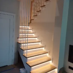 Nowoczesne schody z drewnianymi stopniami podświetlonymi LED, białe pionowe balustrady z prętów, drewniana poręcz, widok z dołu na pierwsze stopnie.