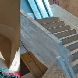 Jasne, wykończone schody z szarobłękitnymi stopniami, białą listwą przypodłogową i balustradą zabezpieczoną folią, widok na ścianę z wzorzystą tapetą i fragment pomieszczenia z zaokrąglonym...