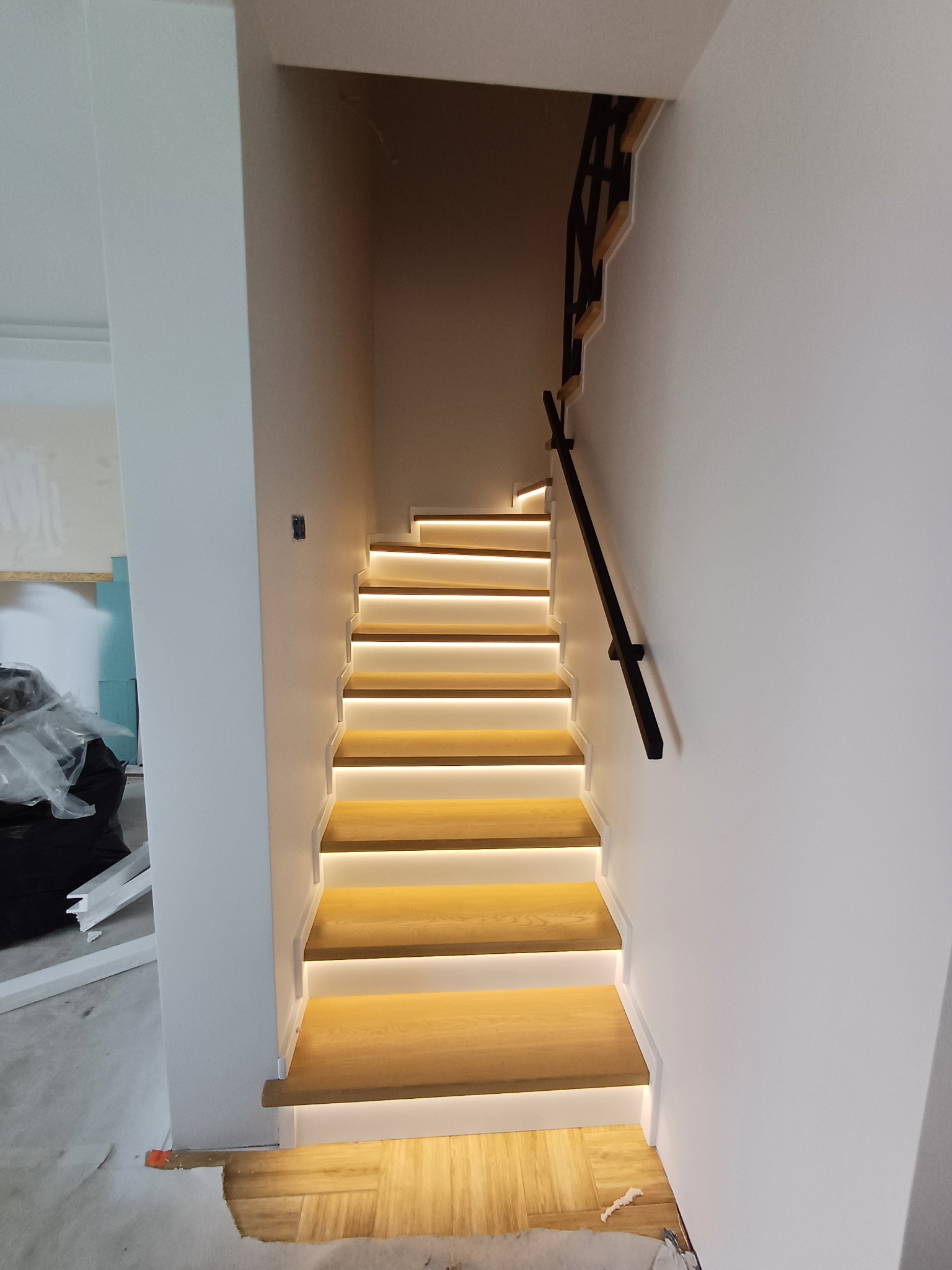 Nowoczesne schody drewniane z podświetleniem LED, czarna poręcz, widok z dołu na górę, fragment pomieszczenia w trakcie wykańczania.