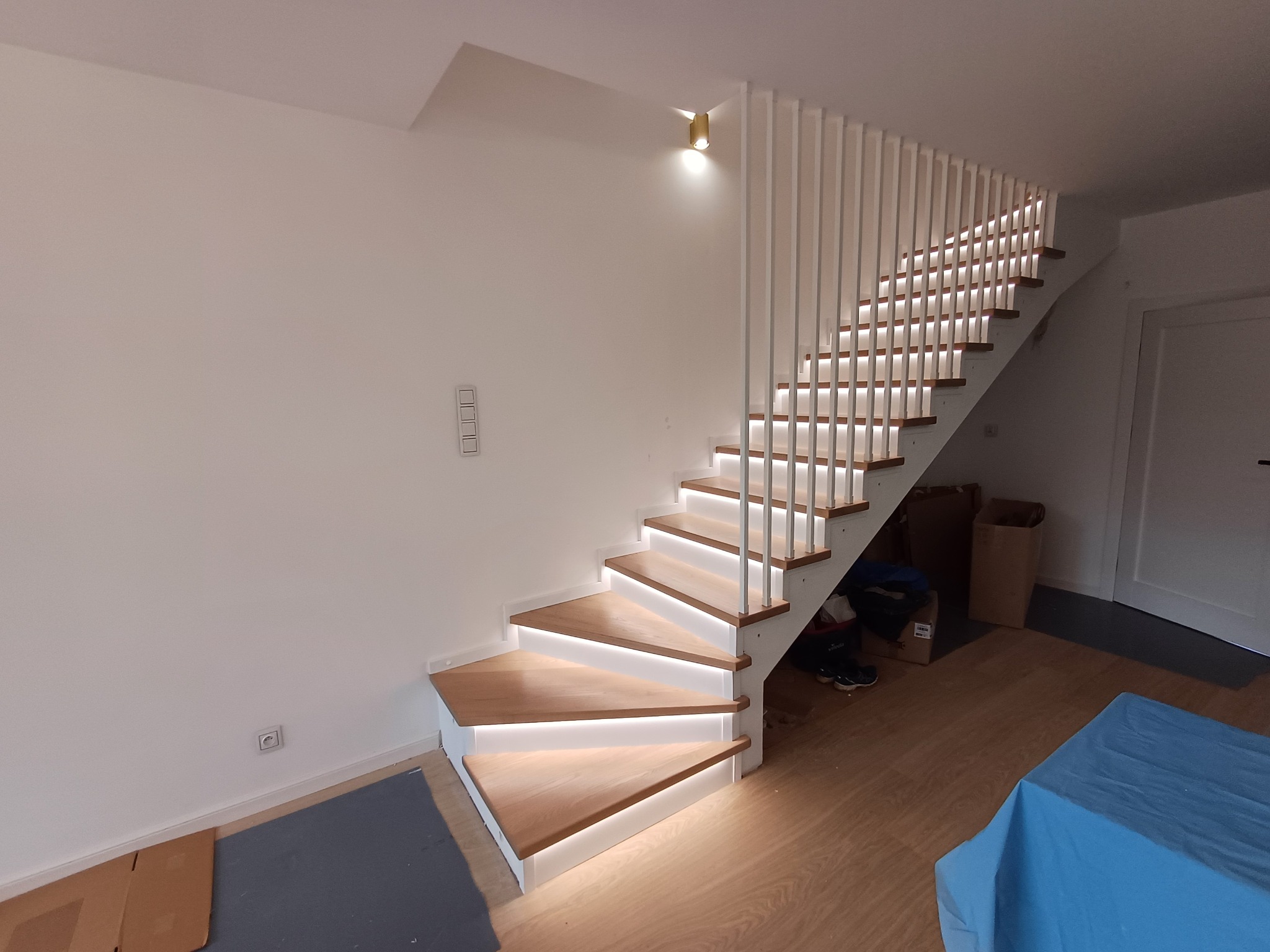 Nowoczesne schody z podświetleniem LED stopni i białymi, pionowymi balustradami, widok z dołu, z fragmentem niebieskiego materiału w dolnym prawym rogu.