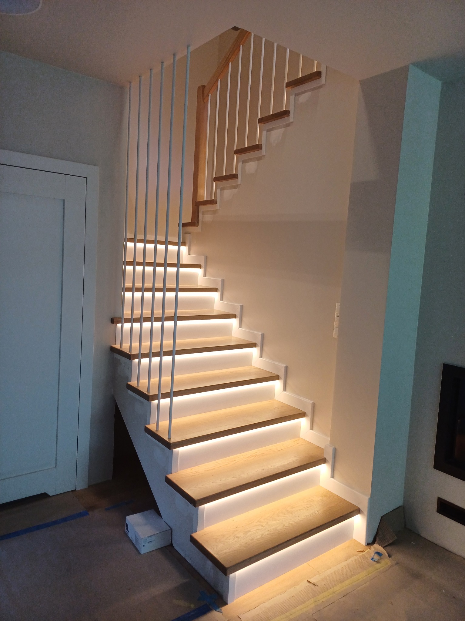Nowoczesne schody z drewnianymi stopniami podświetlonymi LED, białe pionowe balustrady z prętów, drewniana poręcz, widok z dołu na pierwsze stopnie.