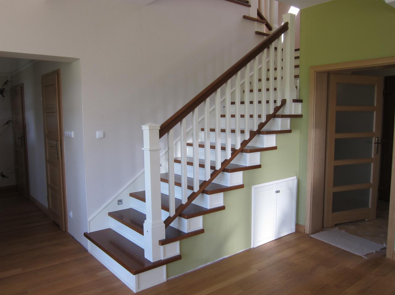 Klasyczne drewniane schody z białymi balustradami i brązowymi stopniami w jasnym wnętrzu domu, widoczne drzwi i podłoga z drewna.