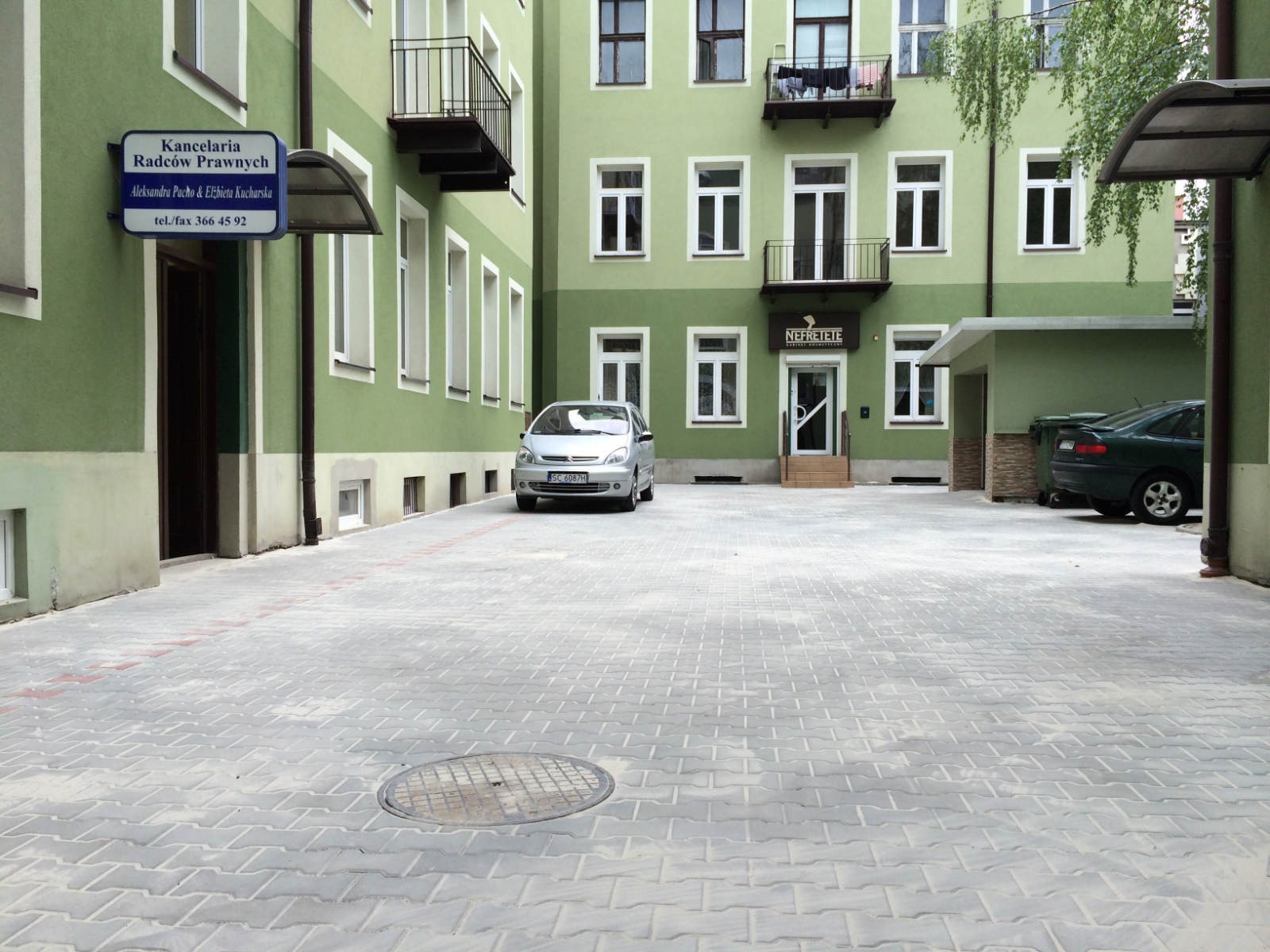 Szara kostka brukowa ułożona na parkingu przed zielonym budynkiem z biurami, widoczny srebrny samochód i studzienka kanalizacyjna na pierwszym planie.