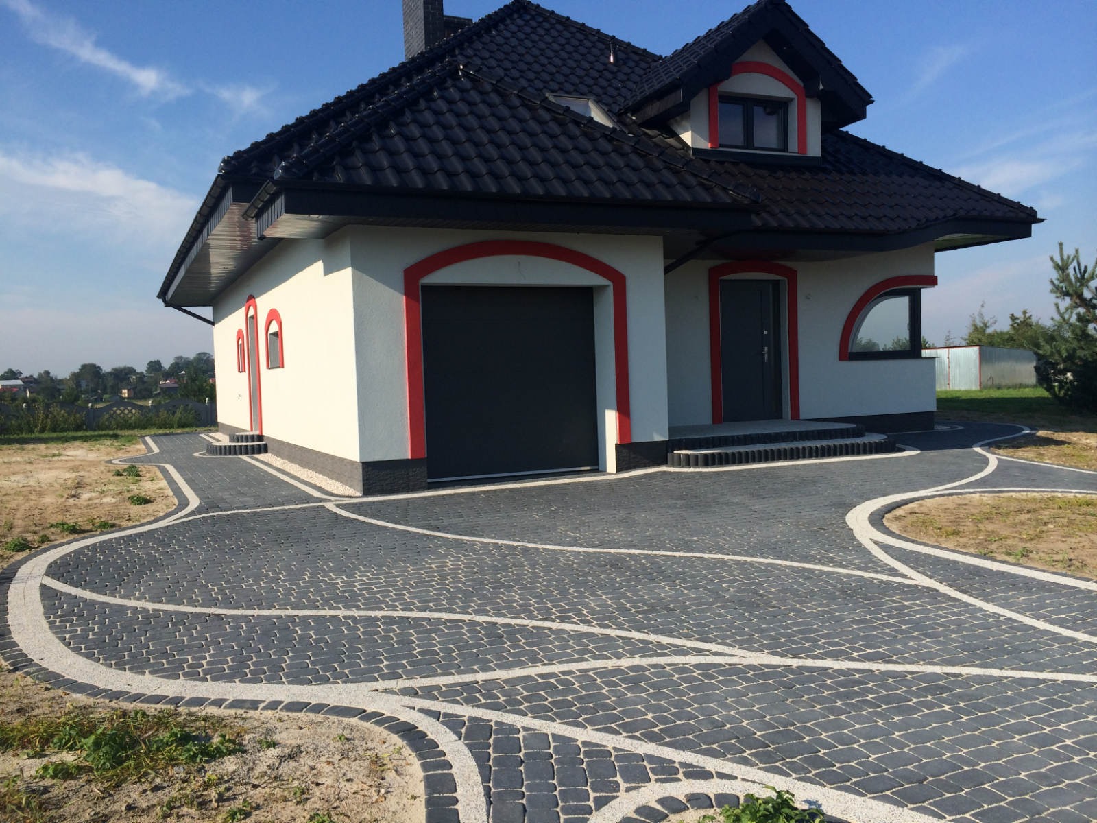 Nowoczesny dom jednorodzinny z czarną dachówką i szarą kostką brukową ułożoną w ozdobny wzór z jasnymi obwódkami.