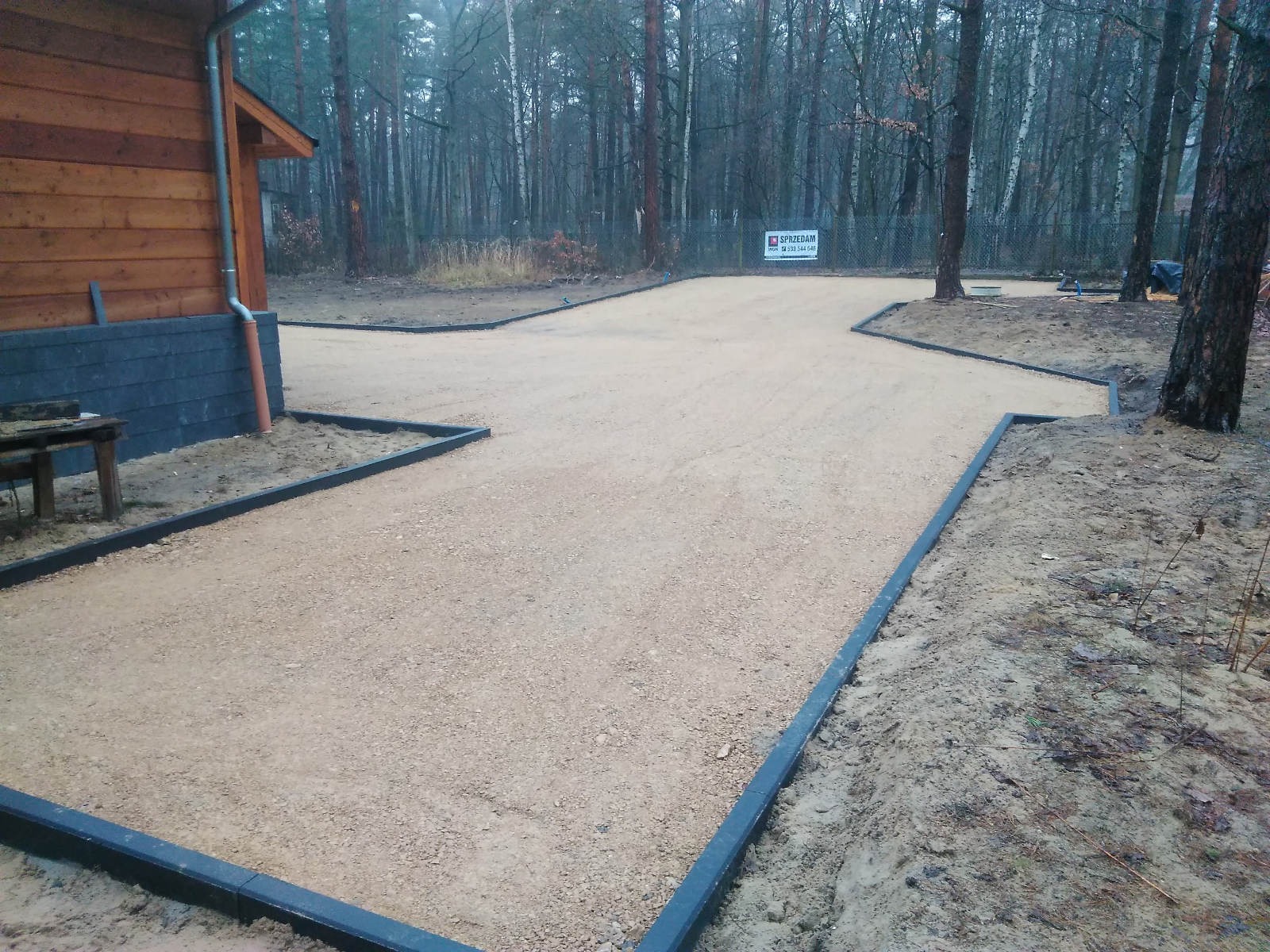 Przygotowany podjazd z obrzeżami z kostki brukowej na działce z drzewami w tle i fragmentem drewnianego budynku.