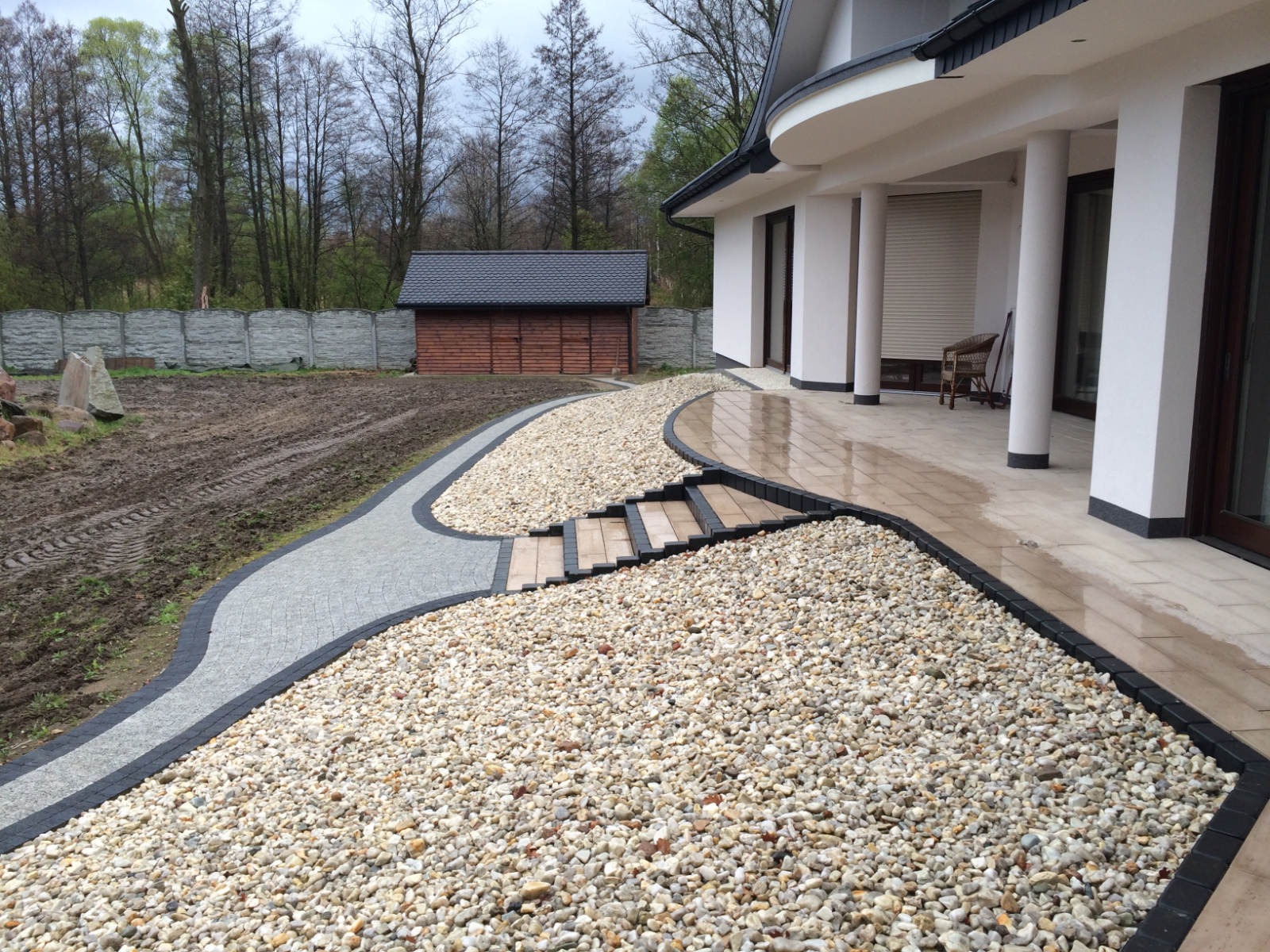 Wykończony teren wokół domu w Częstochowie: ścieżka z kostki brukowej, wysypane kamyki, taras z płyt oraz przygotowany teren pod trawnik z widocznym budynkiem gospodarczym w tle.