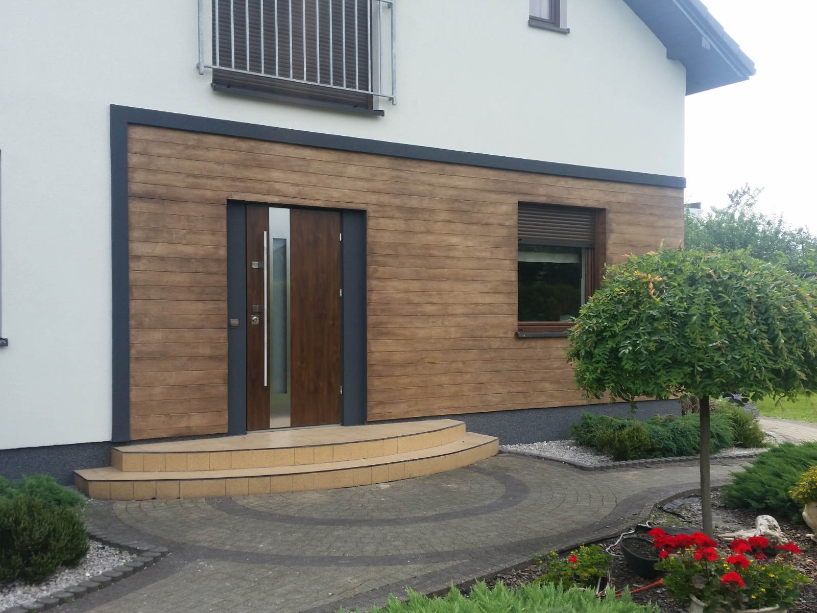 Elewacja domu z drewnianą okładziną, brązowe drzwi wejściowe z elementami stali nierdzewnej, schody z płytek ceramicznych i ścieżka z kostki brukowej w kształcie koncentrycznych kręgów.