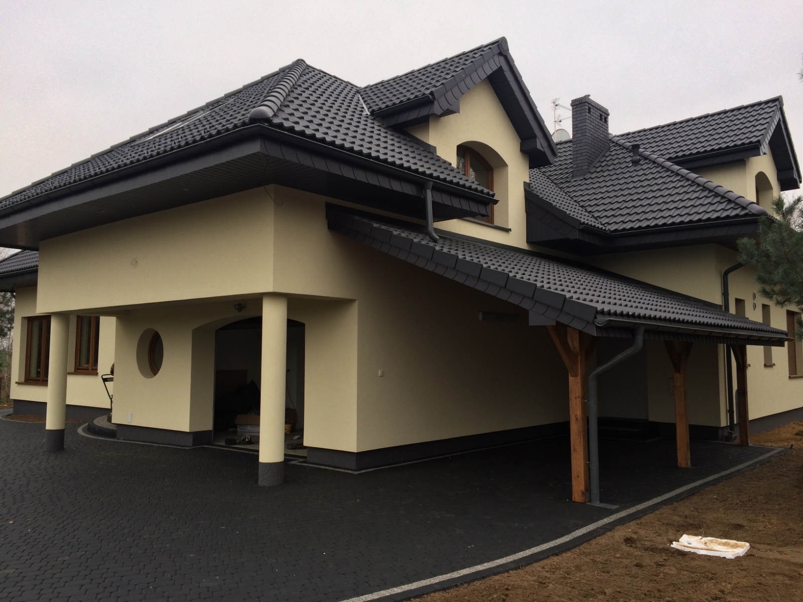 Nowoczesny dom jednorodzinny z czarną dachówką i elewacją w odcieniach beżu, z widocznym fragmentem podjazdu wyłożonego ciemną kostką brukową, sfotografowany w pochmurny dzień.
