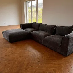 Nowoczesna, narożna sofa w odcieniach szarości z poduszkami, ustawiona na drewnianej podłodze w jodełkę w jasnym, minimalistycznym wnętrzu z dużymi oknami.