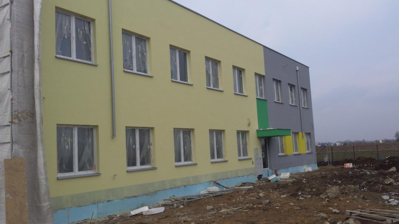 Dwukolorowa elewacja budynku w trakcie prac wykończeniowych; żółta część z oknami zabezpieczonymi folią, szara część z zielonym akcentem nad wejściem, widoczne rusztowania i materiały budowlane...