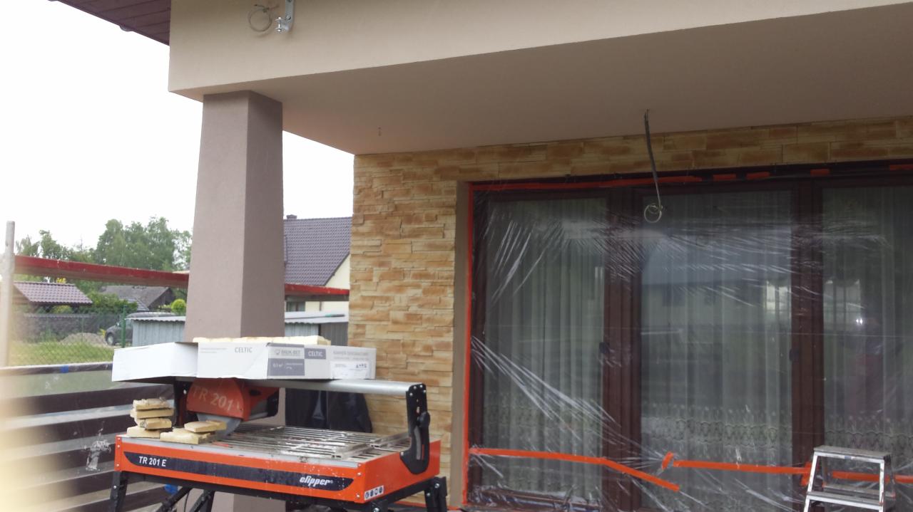 Montaż elewacji z kamienia dekoracyjnego w Piekarach, widoczna maszyna do cięcia płytek i zabezpieczone okno folią.