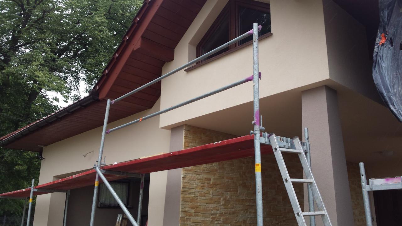 Dom jednorodzinny z beżową elewacją i fragmentem ocieplenia z imitacji kamienia, widoczne rusztowanie z fioletowymi zabezpieczeniami, brązowe okno trójkątne na poddaszu.