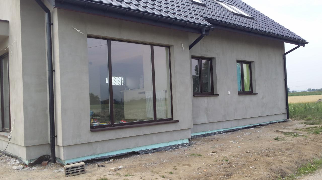 Surowa elewacja domu jednorodzinnego z brązowymi oknami przed ostatecznym wykończeniem, widoczne fragmenty ocieplenia fundamentów i niezabezpieczony teren wokół budynku.