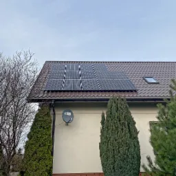 Panele fotowoltaiczne na brązowym dachu domu jednorodzinnego z oknem dachowym, antena satelitarna na elewacji, iglaki w ogrodzie, pochmurne niebo.