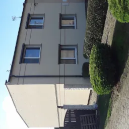 Dwupiętrowy dom z beżową elewacją, widoczne okna z brązowymi ramami, ciemny dach, chodnik z szarej kostki brukowej i ozdobne krzewy.