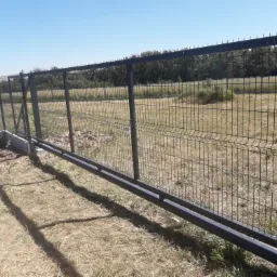 Przesuwna brama z paneli ogrodzeniowych z metalową ramą i betonową podstawą, widok na łąkę i las w tle.