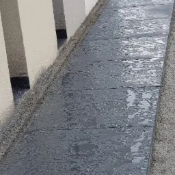 Świeżo ułożona, mokra, ciemnoszara nawierzchnia z prostokątnych płyt, obramowana piaskiem, przylegająca do jasnej ściany budynku.