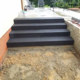 Czterostopniowe schody zewnętrzne z ciemnych płyt kamiennych w trakcie budowy, widoczny wykop i przygotowane podłoże.