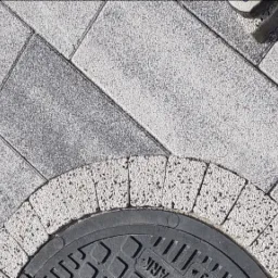 Szara kostka brukowa ułożona w regularny wzór z ciemniejszymi i jaśniejszymi odcieniami, z czarną studzienką kanalizacyjną otoczoną łukiem z jasnej kostki, widoczny fragment buta w prawym górnym rogu.