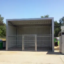 Stalowa wiata z kratą jako ogrodzeniem, przeznaczona na pojemniki na śmieci, widok z zewnątrz przy słonecznej pogodzie.