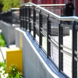 Metalowa balustrada z siatkowym wypełnieniem i stalowymi poręczami na betonowym murku, oświetlona słońcem, z widocznymi białymi tulipanami i żółtą skrzynką w tle.