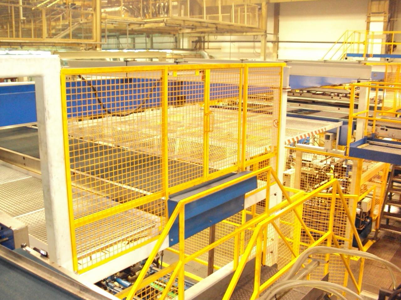 Żółte metalowe barierki ochronne z siatki, zabezpieczające dojście do linii produkcyjnej w hali przemysłowej.