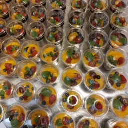 Deser owocowy w przezroczystych pojemnikach z pokrywkami, zawierający mango, maliny, borówki, winogrona i listek mięty, ułożony rzędami na metalowej powierzchni.