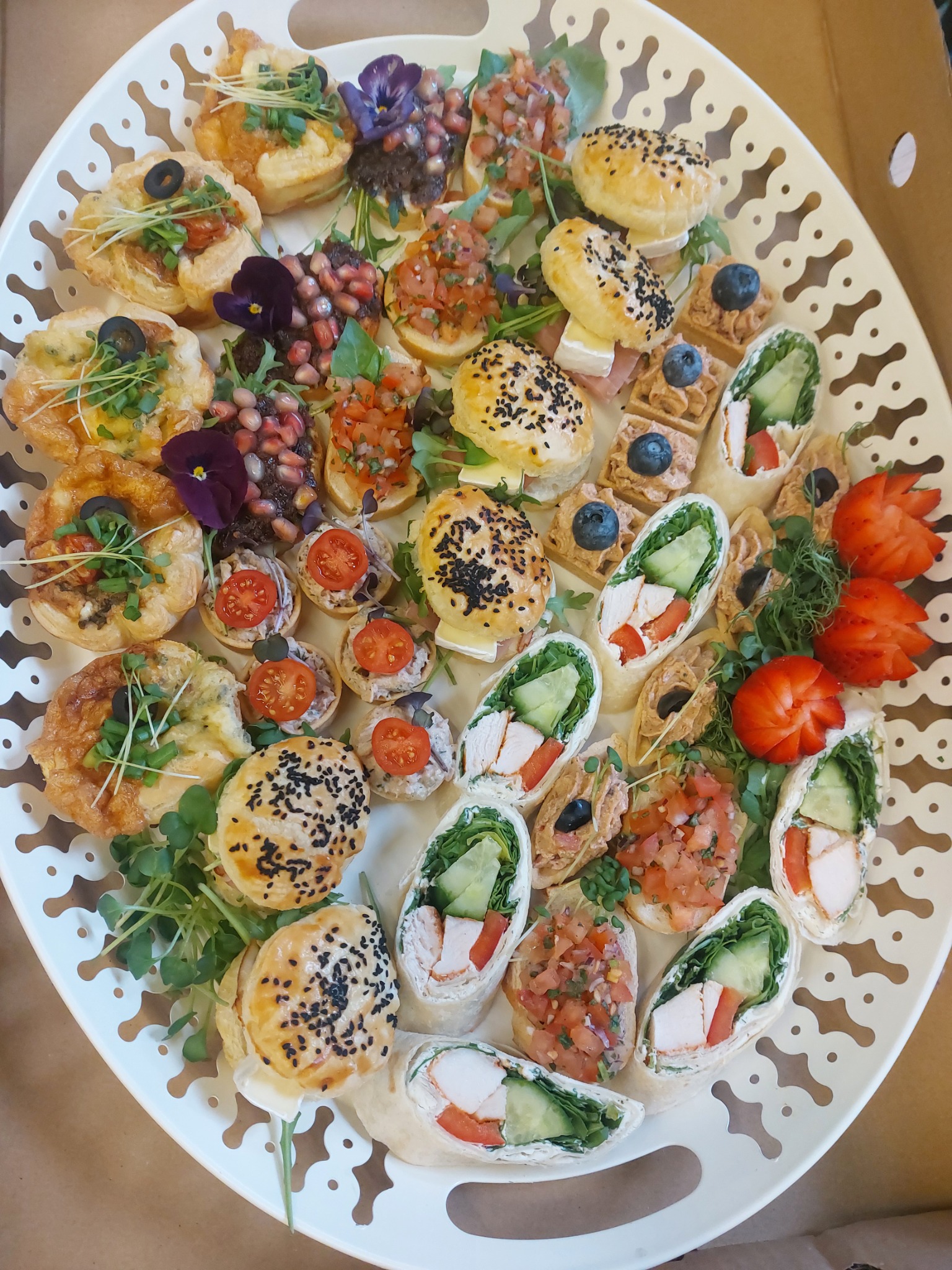Elegancka taca z różnorodnymi przekąskami: mini tartaletki z warzywami i ziołami, bruschetta z pomidorami, roladki z kurczakiem i warzywami, oraz mini ciastka z sezamem, udekorowane kwiatami...