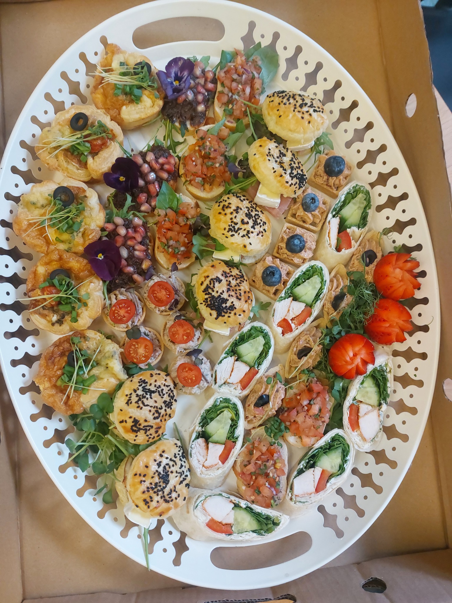 Owalna, biała taca z różnorodnymi, apetycznie wyglądającymi przekąskami: tartaletki z warzywami i oliwkami, mini-kanapeczki z granatem i kwiatami jadalnymi, roladki z kurczakiem i warzywami...