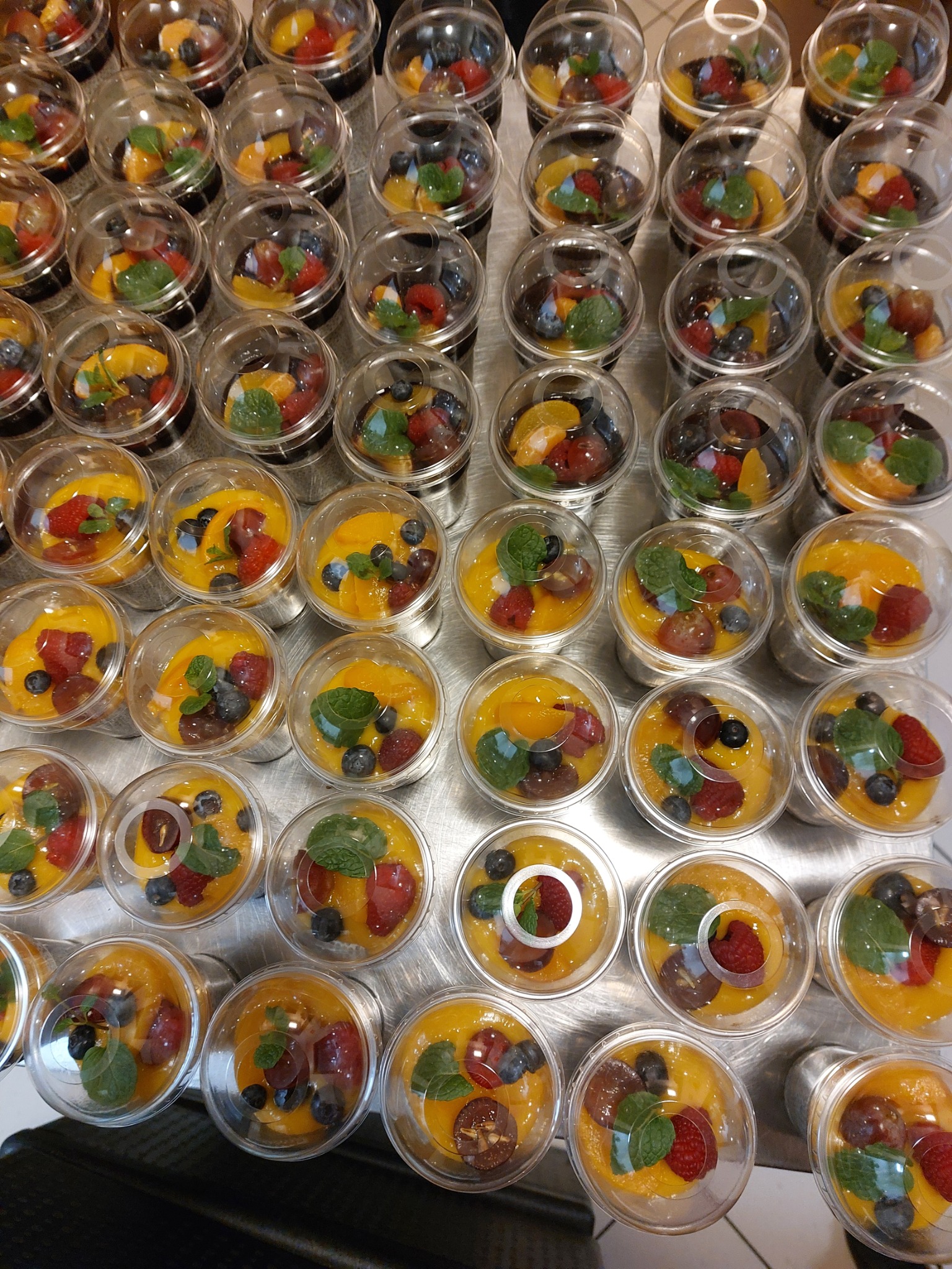 Deser owocowy w przezroczystych pojemnikach z pokrywkami, zawierający mango, maliny, borówki, winogrona i listek mięty, ułożony rzędami na metalowej powierzchni.