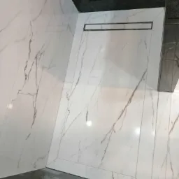 Wykończona łazienka z białymi marmurkowymi płytkami na ścianach i podłodze, czarny odpływ liniowy, widok z góry.