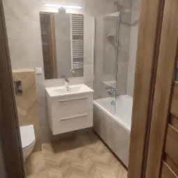 Nowoczesna łazienka z szarymi płytkami imitującymi marmur, białą szafką umywalkową z dwiema szufladami, lustrem z oświetleniem LED, kabiną prysznicową nad wanną i podłogą w jodełkę.