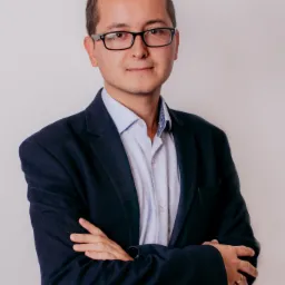 Portret mężczyzny w okularach i granatowej marynarce, skrzyżowane ręce, białe tło.