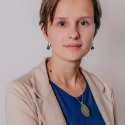 Portret kobiety z krótkimi brązowymi włosami, ubranej w beżową marynarkę i niebieską bluzkę, z delikatną srebrną biżuterią i kolczykami z ametystami, na jasnym tle.