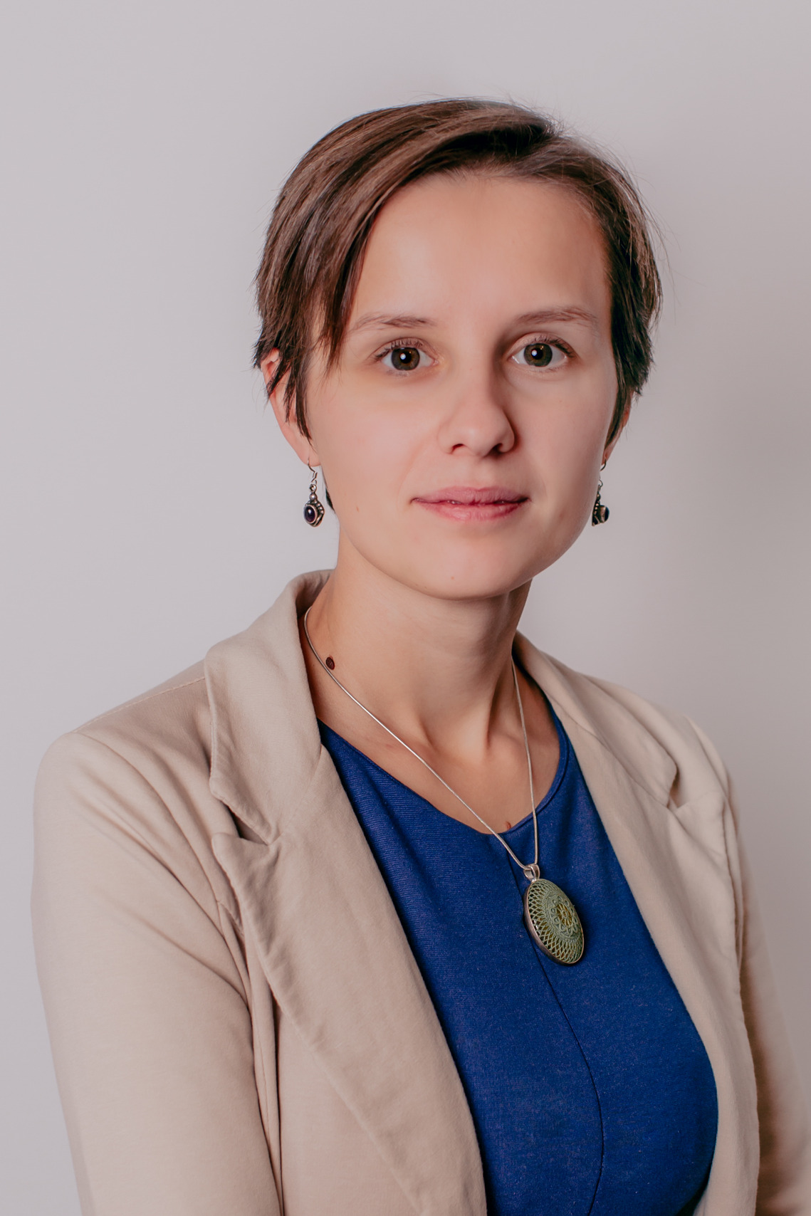 Portret kobiety z krótkimi brązowymi włosami, ubranej w beżową marynarkę i niebieską bluzkę, z delikatną srebrną biżuterią i kolczykami z ametystami, na jasnym tle.